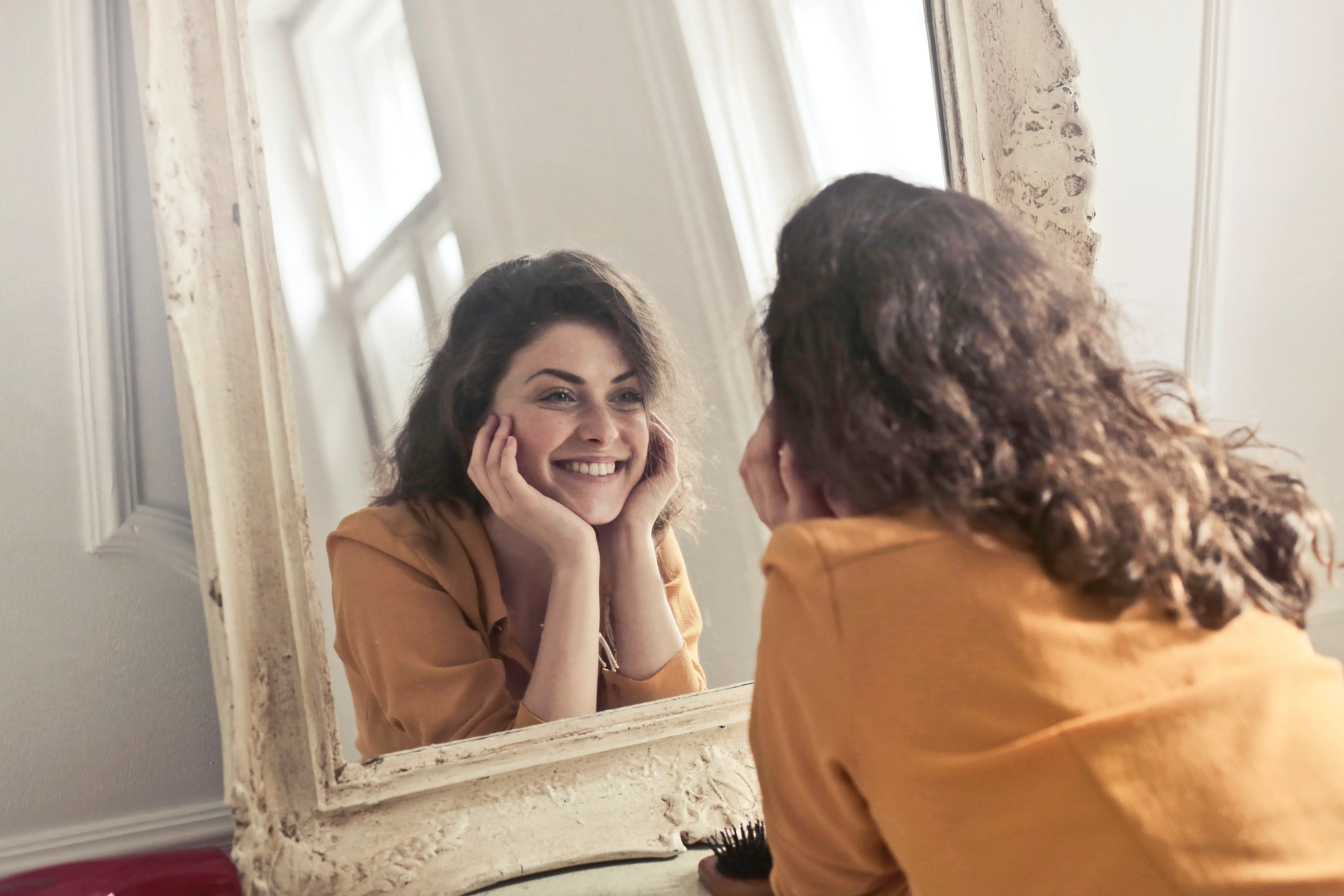 A brown-haired woman looks into the mirror and smiles at her reflection