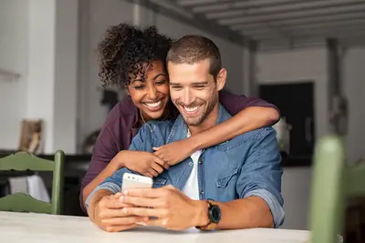 A woman hugs her man who smiles at his phone